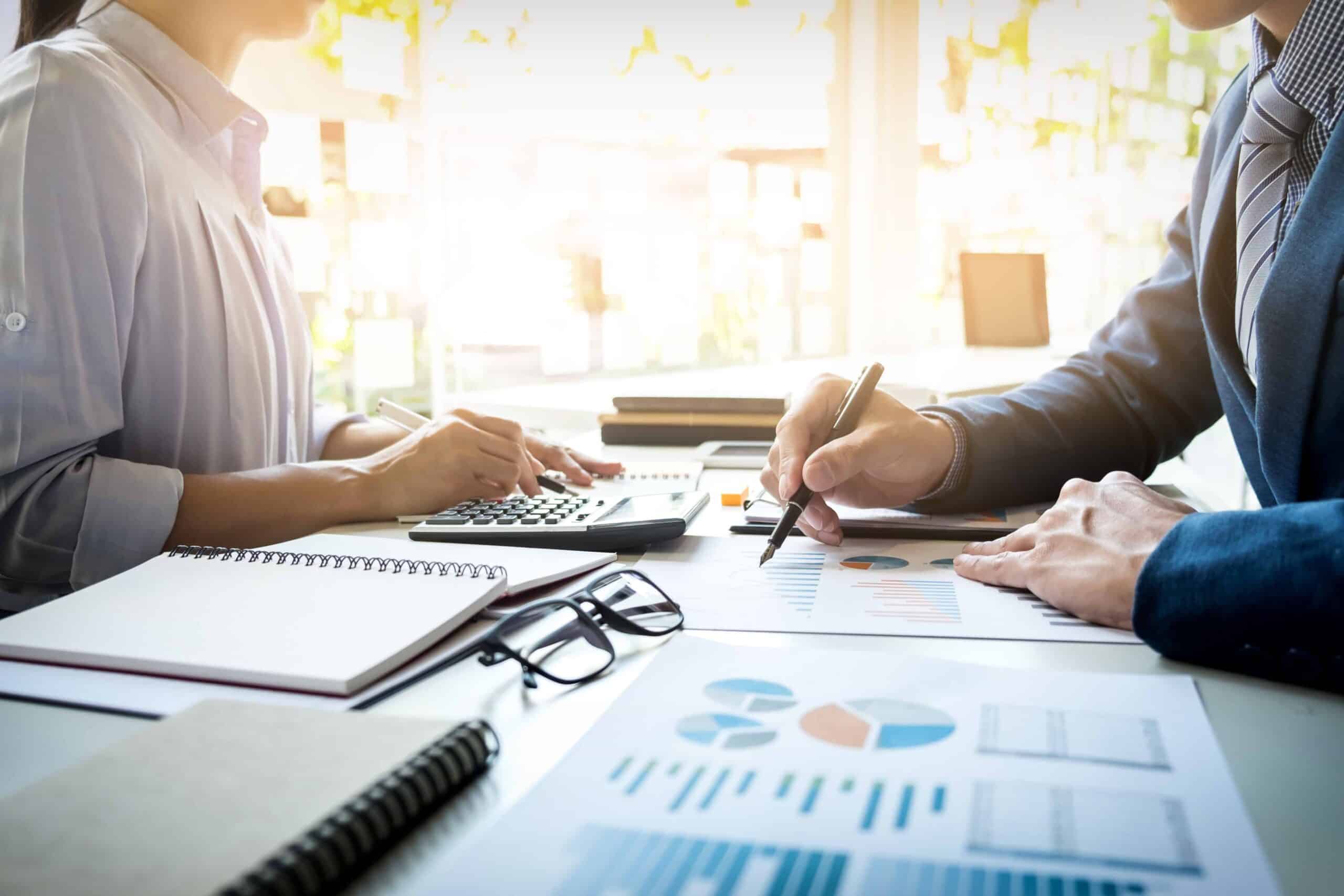 Two professionals analyze bookkeeping data at a desk, one using a calculator while the other writes on graphs and charts. Notebooks and glasses nearby.