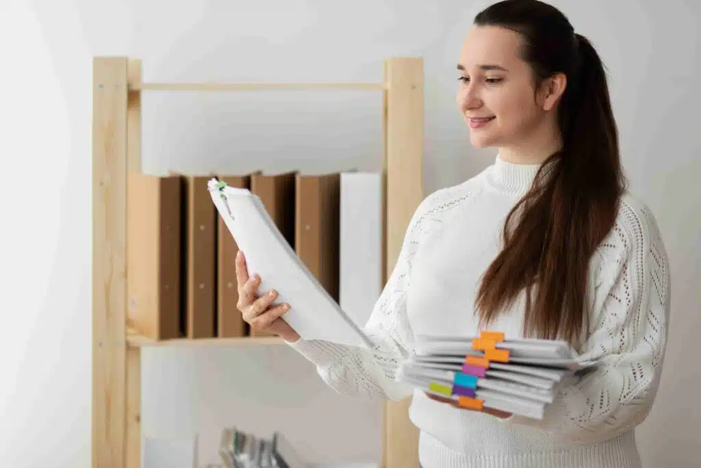 A person in a white sweater holds a clipboard and a stack of organized documents for bookkeeping, standing amid a wooden shelf with file boxes.